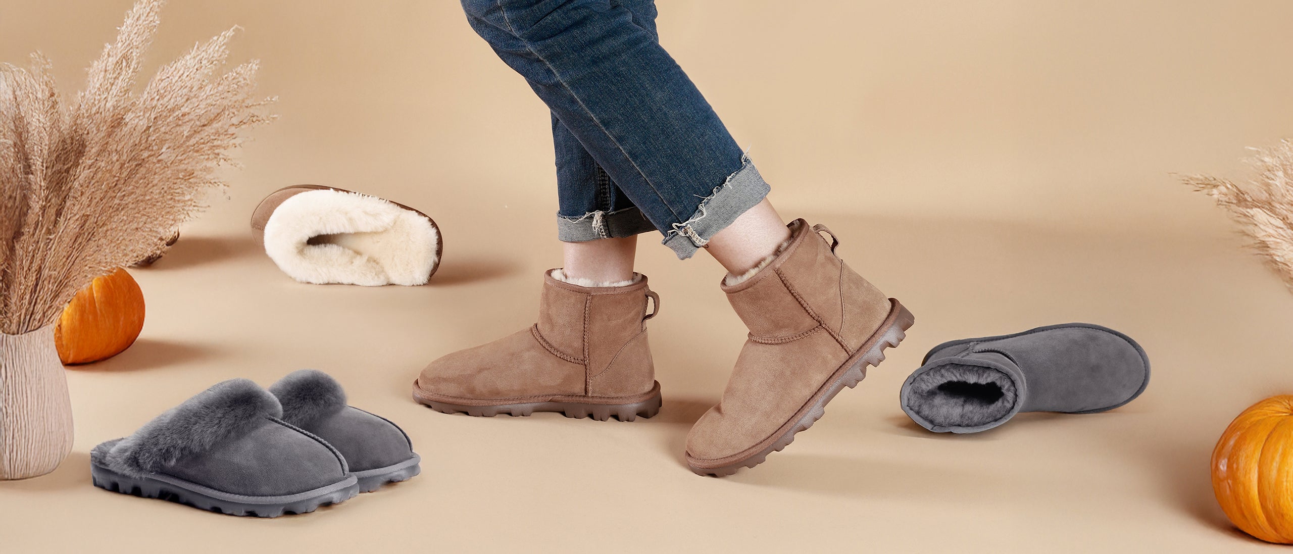 A woman wearing a pair of Cozie Steps brown shearling boots with gray and brown slippers lying on a beige background surrounded by autumn decorations